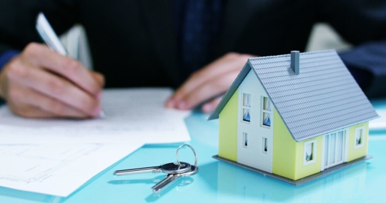 Small house and keys on table with person signing papers in background.